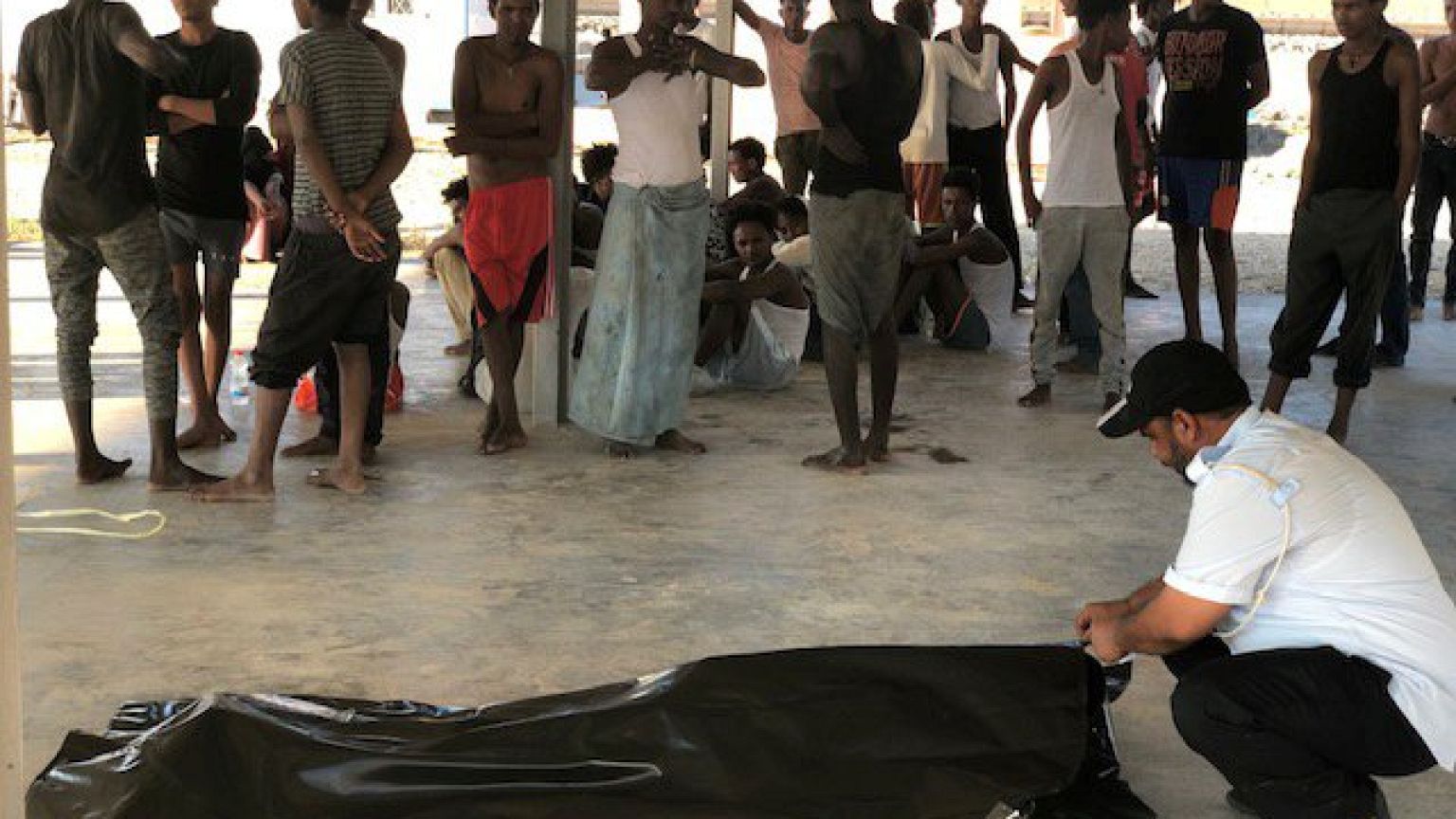 Image: Migrants watch the body of their fellow migrant who died after a woo