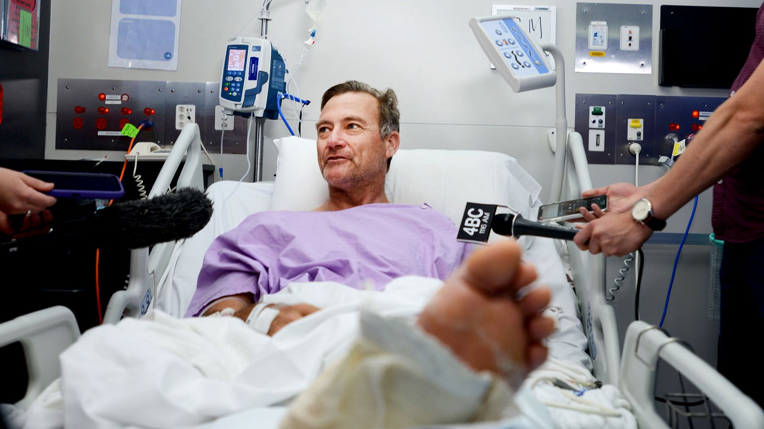 Image: Neil Parker speaks to reporters from his hospital bed while recoveri