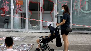 Image: A woman with a stroller stands in front of the smashed glass door of