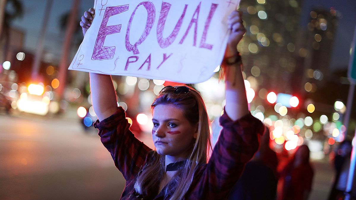 Image: A woman holds a sign that reads, "Equal Pay" during "A Day Without A