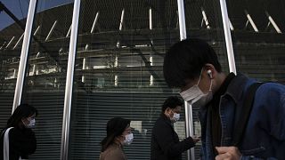 Image: People with masks walk past a building reflecting the New National S