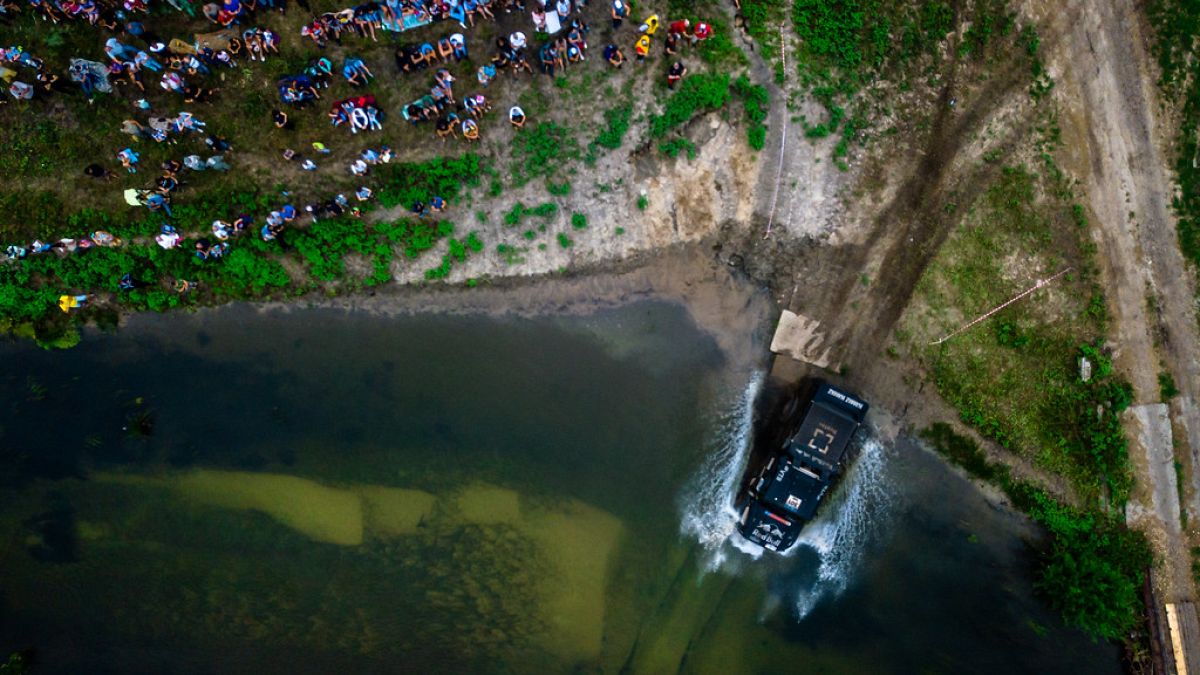 Vogelperspektive: Ein Truck fährt in ein Gewässer, viele Menschen schauen