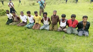 Rohingya men kneel with hands bound as a paramilitary officer stands guard.