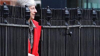 Theresa May in rotem Mantel zwischen schwarzem Zaun