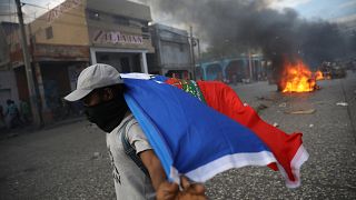 Ein vermummter Mann trägt eine Haiti-Flagge, Im Hintergrund brennen Reifen
