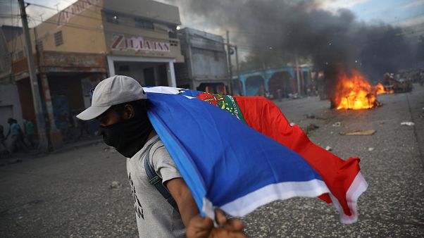 Ein vermummter Mann trägt eine Haiti-Flagge, Im Hintergrund brennen Reifen Ein vermummter Mann trägt eine Haiti-Flagge, Im Hintergrund brennen Reifen