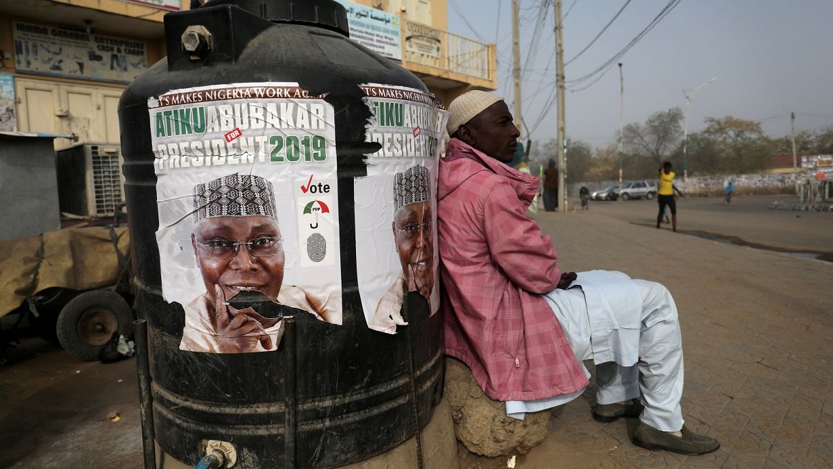 A man sits next to a campaign poster after Nigeria's elections are delayed.