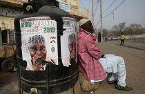 A man sits next to a campaign poster after Nigeria's elections are delayed.