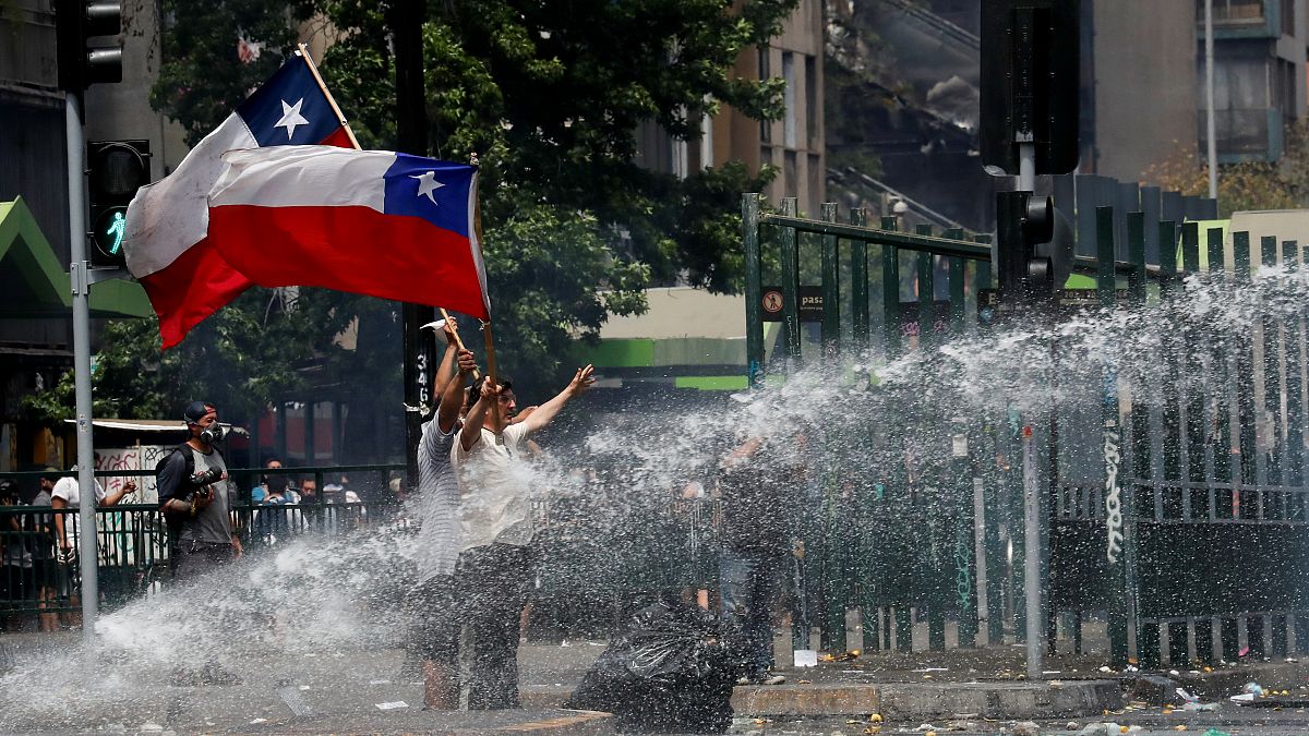 المتظاهرون يلوحون بالأعلام التشيلية بينما تقوم قوات الأمن برشهم بخراطيم المياه أثناء الاحتجاج على النموذج الاقتصادي للدولة في تشيلي في سانتياغو، 23 أكتوبر / تشرين الأول 2019