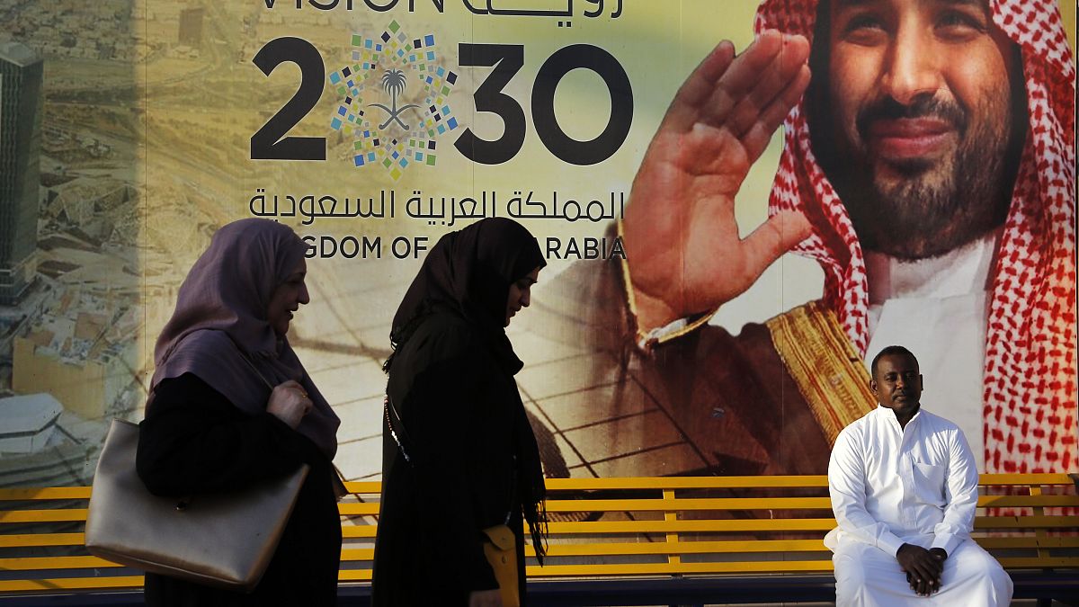 سعوديون أمام لافتة لولي العهد السعودي محمد بن سلمان ورؤية 2030، في مركز تجاري في جدة، المملكة العربية السعودية.