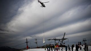People look at an helicopter carrying snow in order to place it on a ski slope in the Superbagneres station in French Pyrenees mountain, on February 15, 2020.