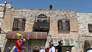 جندي إسرائيلي يراقب من أعلى سطح منزل استعراض "البوريم" عبر شارع الشهداء، وعائلة فلسطينية تراقب من شباك منزلها الاستعراض.