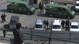 People, some wearing protective face masks and suits as a preventive measure against the spread of the coronavirus wait gather around hearse trucks carrying coffins of victims