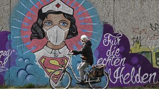 سيدة ألمانية تسير بدراجتها بمدينة هام أمام جدارية كبيرة يظهر الممرضة كالمرأة العجيبة، وكتب عليها "لأجل الأبطال الحقيقيين". 13/04/2020