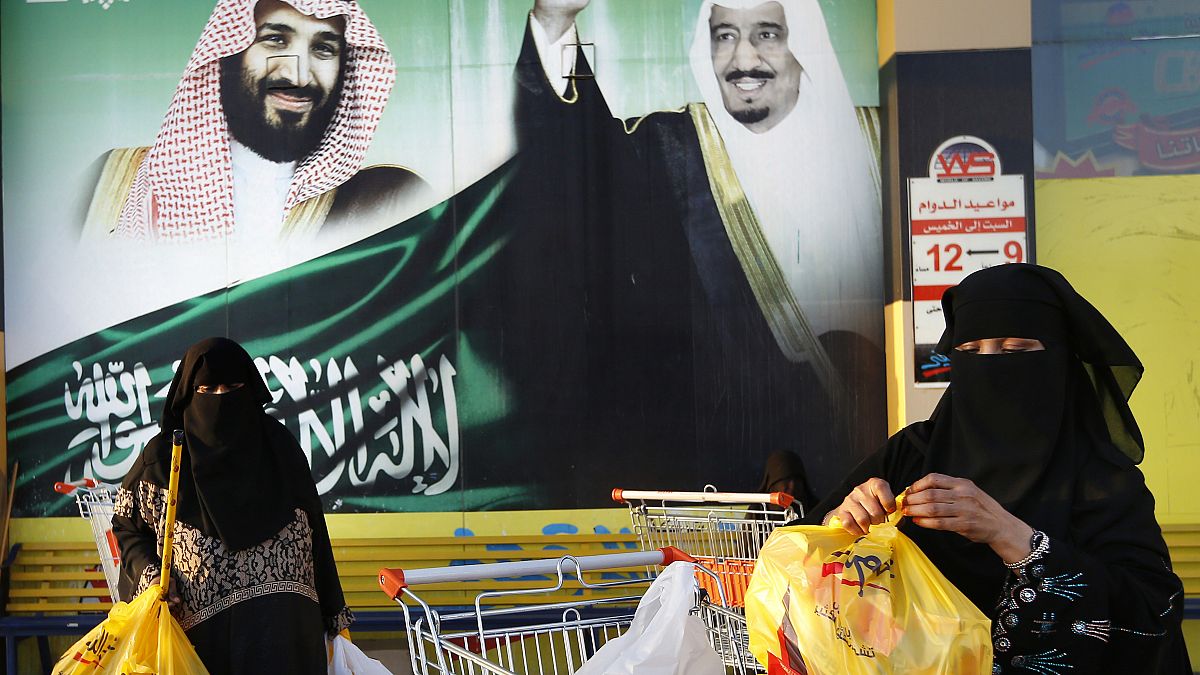 نساء تحملن أكياس التسوق أمام لافتة تظهر الملك السعودي سلمان وولي عهده الأمير محمد بن سلمان، خارج مركز تجاري في جدة.