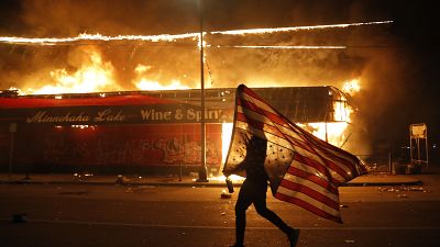 Un hombre con una bandera estadounidense frente a un comercio en llamas en Mineápolis