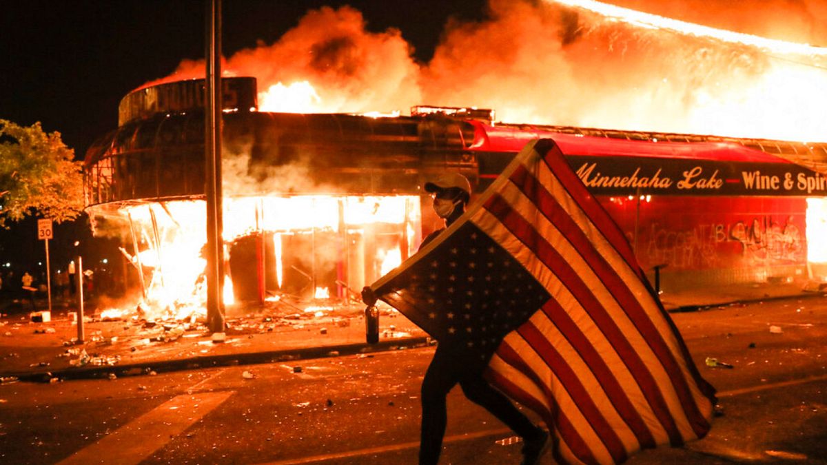 A protester carries a U.S. flag upside, a sign of distress, next to a burning building, early Friday, May 29, 2020, in Minneapolis.
