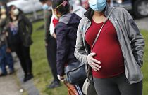 A pregnant woman wearing a face mask and gloves holds her belly as she waits in line for groceries with hundreds during a food pantry.