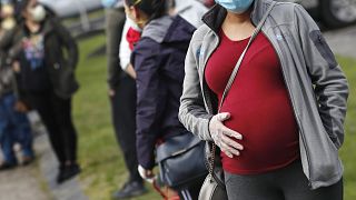 En esta foto de archivo del jueves 7 de mayo de 2020, una mujer embarazada con una mascarilla y guantes se sostiene el vientre mientras espera en la cola para hacer la compra.