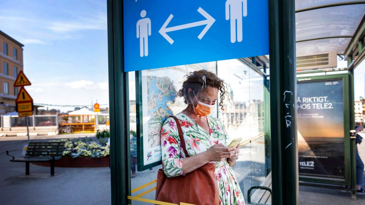 A woman wears a face mask at a bus stop, with an information sign asking people to keep social distance due to the corona pandemic, in Stockholm, Friday, June 26, 2020.