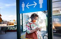 A woman wears a face mask at a bus stop, with an information sign asking people to keep social distance due to the corona pandemic, in Stockholm, Friday, June 26, 2020.