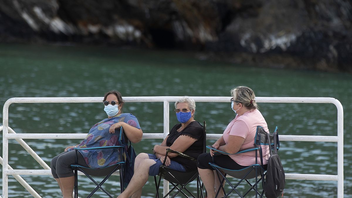 Three women wearing protective masks sit by the dock in Porto o Barqueiro, north western Spain, Sunday, Aug. 2, 2020.