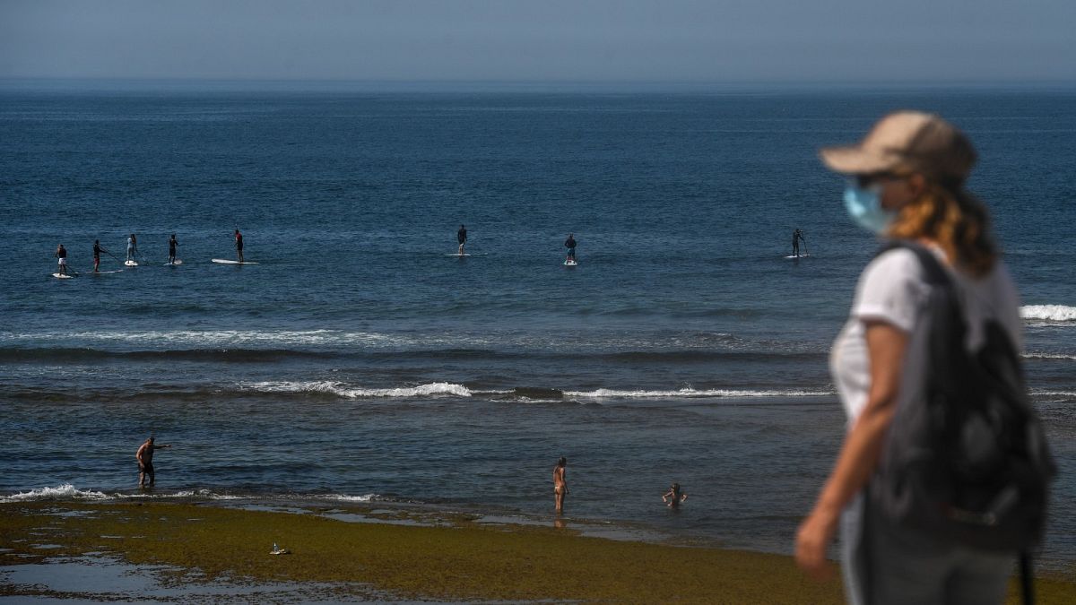 A woman wearing a face mask walks past paddle surfers riding waves at Parede beach in Cascais in the outskirts of Lisbon