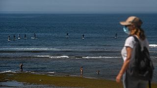 A woman wearing a face mask walks past paddle surfers riding waves at Parede beach in Cascais in the outskirts of Lisbon