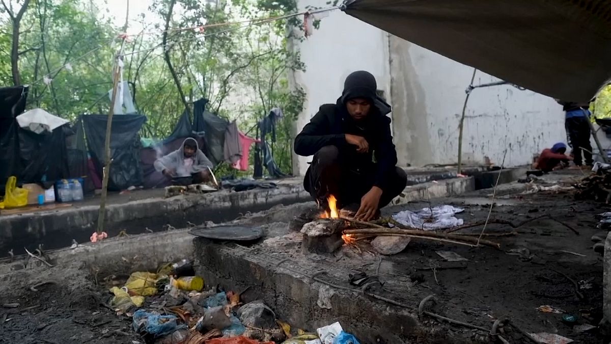 Migrants standing next to makeshift tents inside rundown building on edge of forest