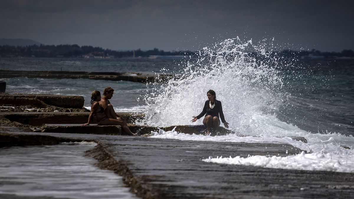 Una mujer habla con dos personas mientras una ola les salpica en La Habana