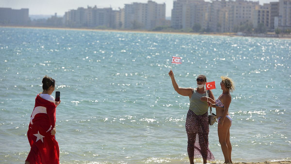 زوار يحملون الأعلام التركية المنفصلة يلتقطون صورًا لبعضهم البعض على الشاطئ مع المباني المهجورة بعد أن فتحت الشرطة شاطئ فاروشا.