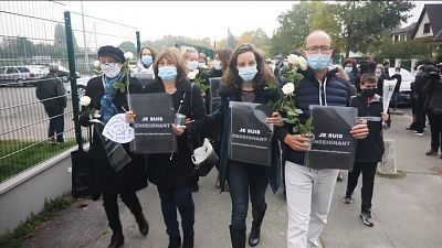 Various of mourners walking with flowers and placards reading (French) 'I am a teacher'
