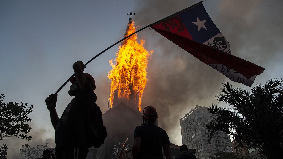 Un manifestante enarbola una bandera de Chile frente a la cúpula de la Asunción en llamas