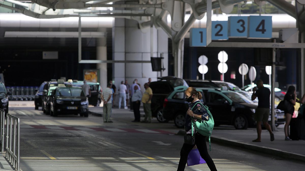 A woman wearing a face mask walks outside Lisbon's airport while in the background taxi drivers stand idle waiting for passengers, Friday, Sept. 11, 2020. Portugal
