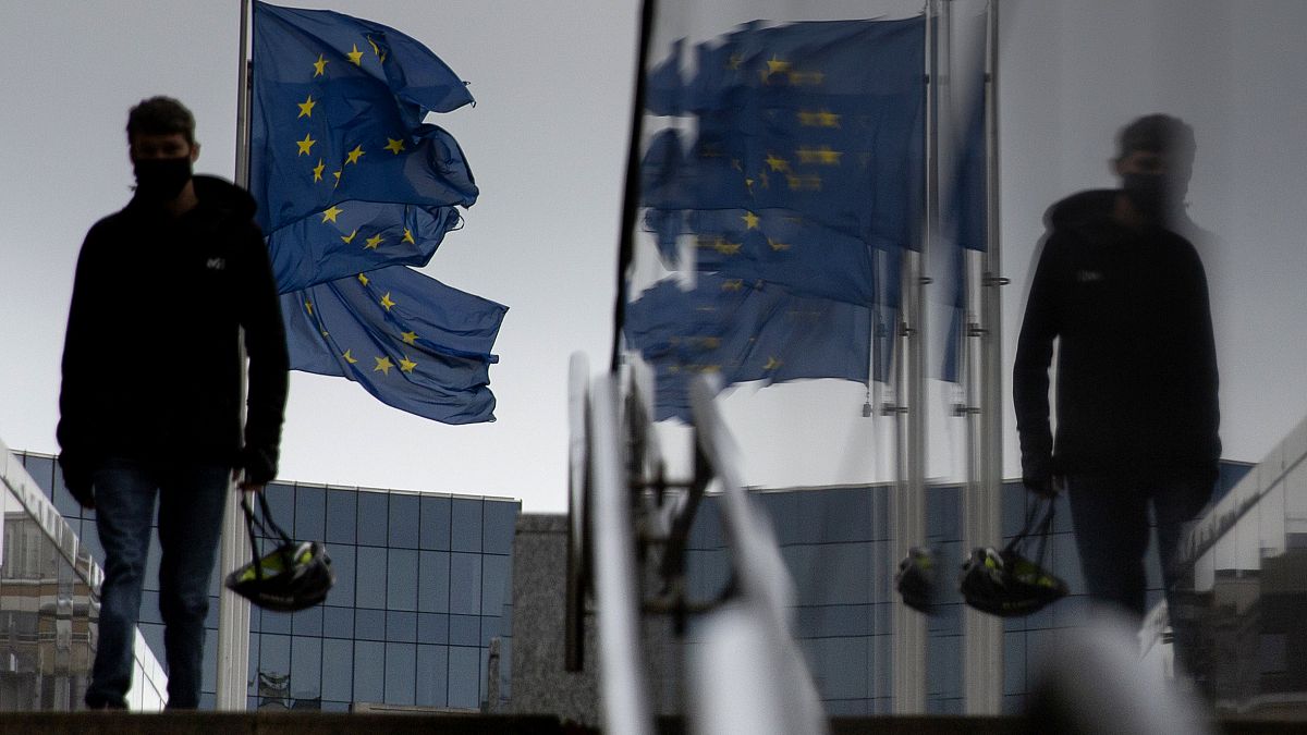 A man carries a cycling helmet as he walks by EU flags outside EU headquarters in Brussels, Wednesday, Oct. 28, 2020.