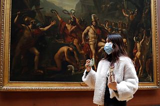 Masked tourist walks by an oil on canvas of 1814 entitled Leonidas at Thermopylae, by Jacques Louis David, at the Louvre Museum in Paris