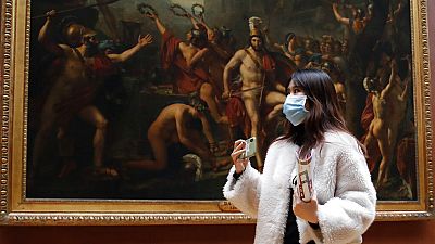 Masked tourist walks by an oil on canvas of 1814 entitled Leonidas at Thermopylae, by Jacques Louis David, at the Louvre Museum in Paris