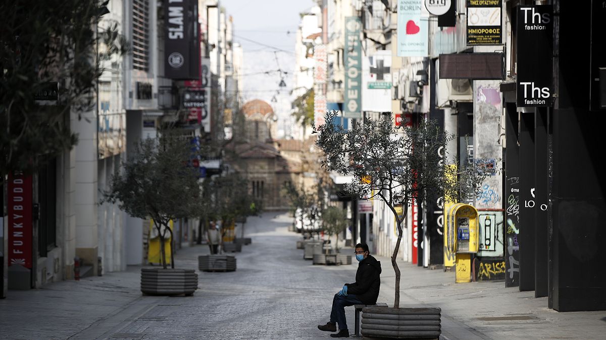 A man wearing a face mask and plastic gloves to protect from coronavirus sits in the empty Ermou Street