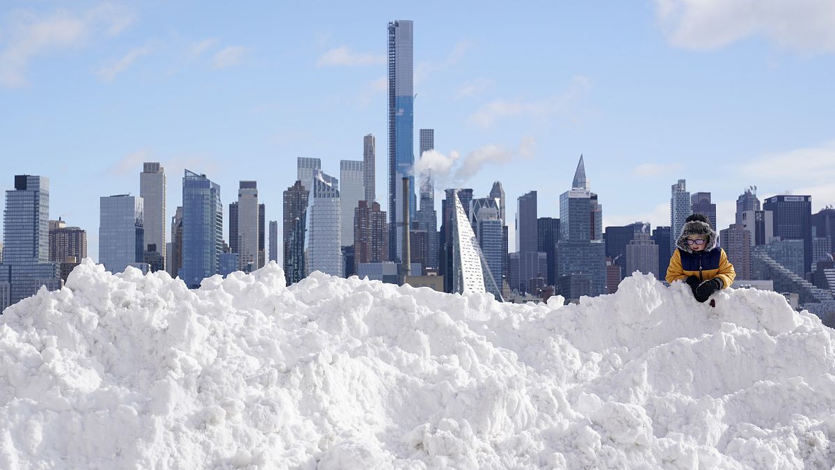 A boy plays on a mound of snow in front of the skyline of New York City 