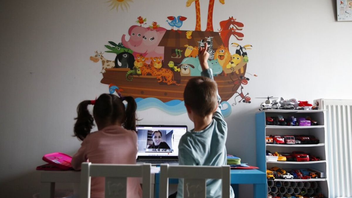 A kindergarten teacher reads a fairy story as Panos raising his hand with his twin sister Marina watch the online lesson in Athens