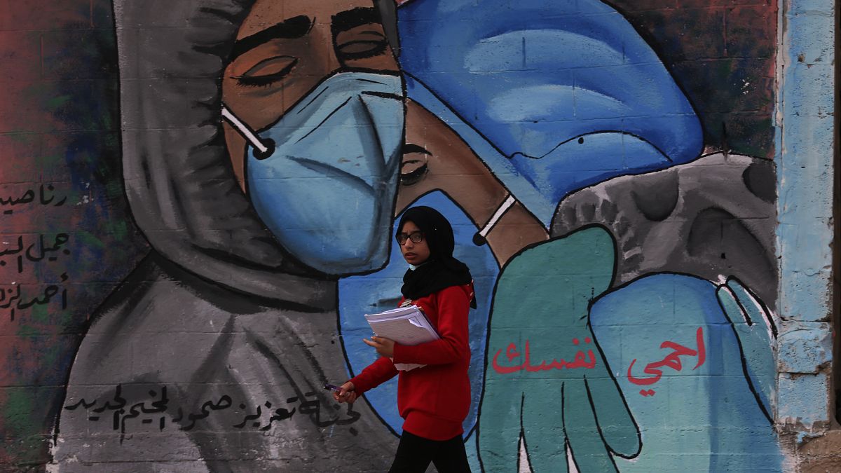 طالبة تمشي أمام لوحة جدارية تشجع على ارتداء كمامة للوقاية من فيروس كورونا، على الطريق الرئيسي لمخيم النصيرات، وسط قطاع غزة. 