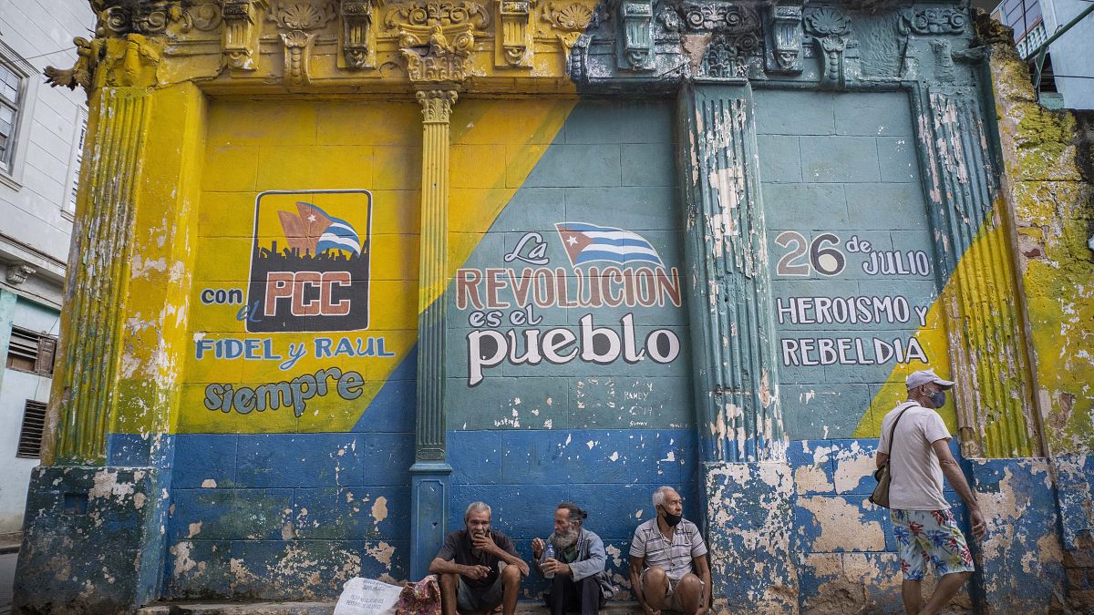 Tres hombres con las mascarillas mal colocadas charlan sentandos delante de un muro con consignas políticas en La Habana