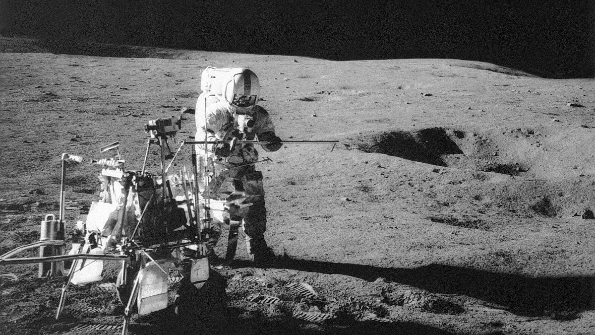 Apollo 14 astronaut Alan B. Shepard Jr. conducts an experiment near a lunar crater, using an instrument from a two-wheeled cart carrying various tools. 