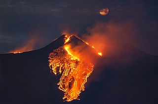 The moon is partially seen in the sky as lava flows from the Mt Etna volcano, near Catania in Sicily, southern Italy, early Tuesday, Feb. 23