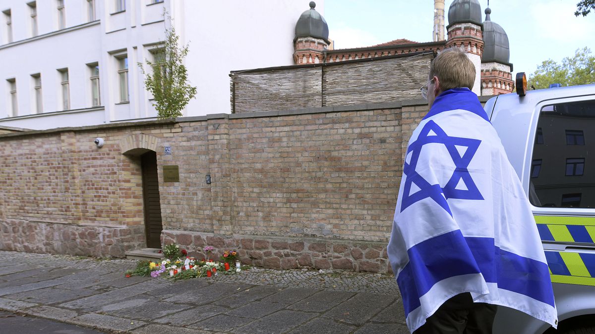 شخص يحمل علم إسرائيل ويقف بجانب زهور وشموع أمام كنيس يهودي في هاله، ألمانيا