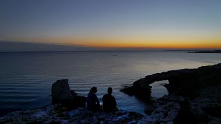 A man and a woman sit near the natural rock bridge that is named "Love Bridge", during sunset at the Ayia Napa