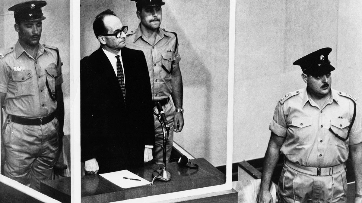 أدولف أيخمان يقف في قفص زجاجي، محاطا بحراس، في قاعة محكمة القدس أثناء محاكمته عام 1961 