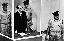 أدولف أيخمان يقف في قفص زجاجي، محاطا بحراس، في قاعة محكمة القدس أثناء محاكمته عام 1961