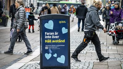 People walk past a bin with a sign reading "The danger is not over. Keep your distance" in a pedestrian street in central Uppsala, Sweden. October 21, 2020. People walk past a bin with a sign reading "The danger is not over. Keep your distance" in a pedestrian street in central Uppsala, Sweden. October 21, 2020.