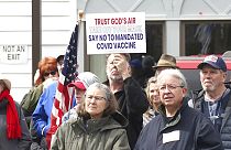A sign denouncing the COVID-19 vaccine is held up in the crowd gathered at a protest